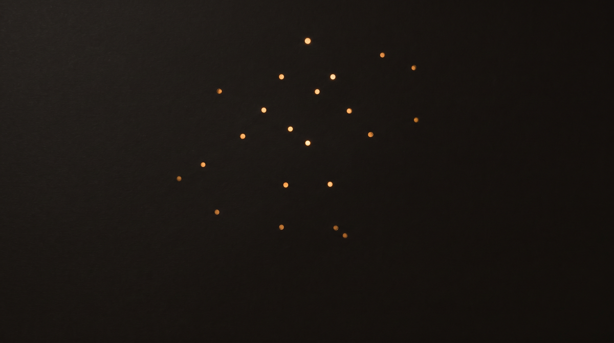 Scattered warm golden light points glowing against a deep black background, resembling stars or fiber-optic lights in a minimalist dark composition