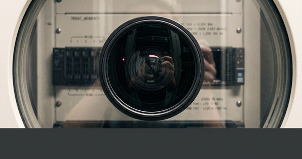 A camera lens reflected in curved glass — one memory module being removed from a server rack visible behind — a single point of red — the removal of what watches back.